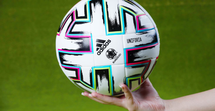 Eine Hand hält einen Fussball von Adidas. Im Hintergrund ist grüner Rasen erkennbar